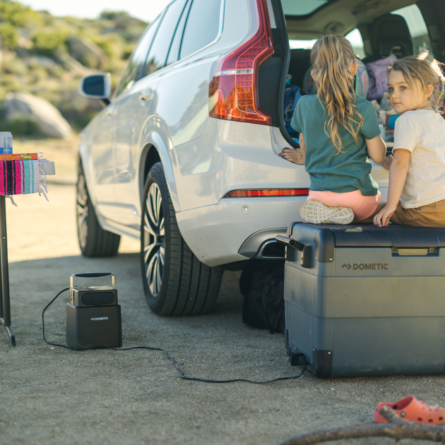 Two children sitting on a cooler next to an SUV with a power station and cooler in the foreground.